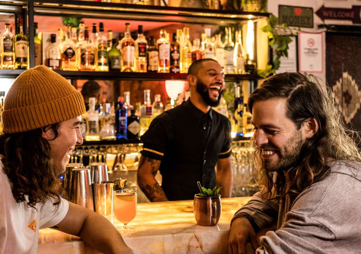 Dos personas jóvenes en la barra de La Destilería con dos cócteles y el camarero de fondo riéndose